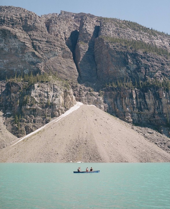 Three people in a canoe paddle across a turquoise lake in front of a large mountain.