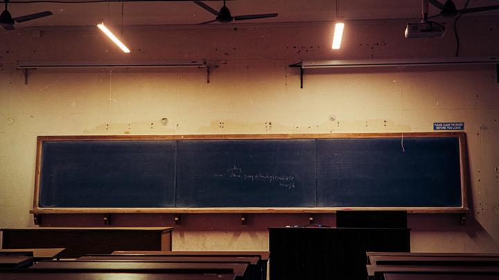 A classroom with a large blackboard, wooden desks, and ceiling fans is illuminated by fluorescent lights.