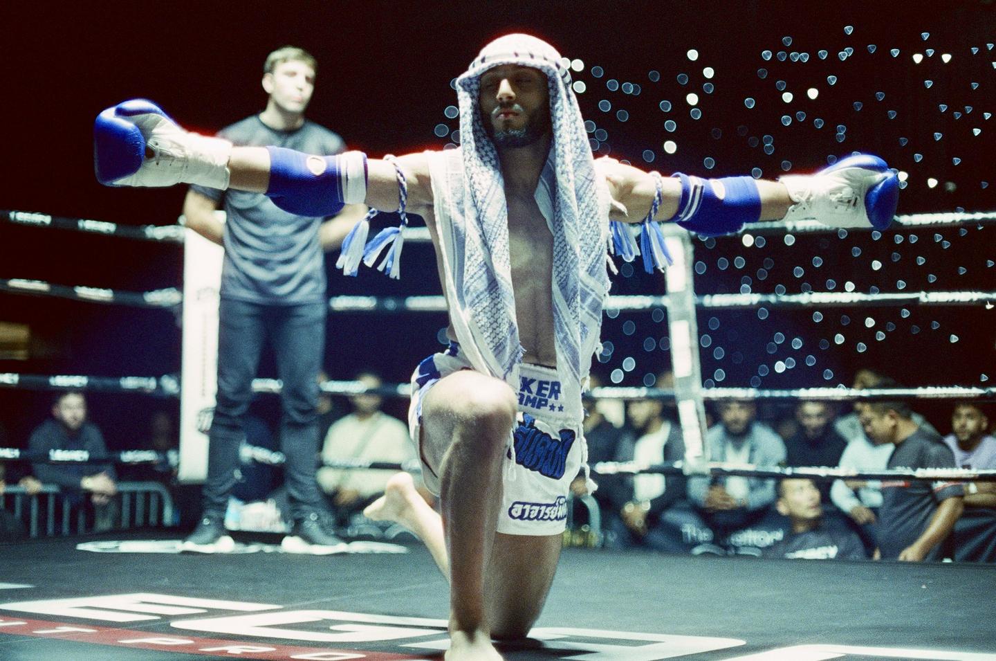 A fighter in a boxing ring kneels with arms outstretched, wearing boxing gloves and a keffiyeh.