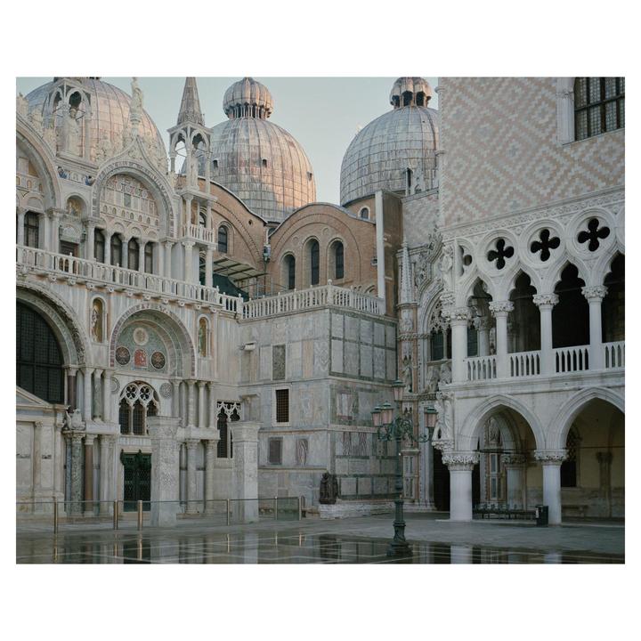 A large ornate building with multiple domes and arches stands under a clear sky, with reflections on the wet ground in the foreground.