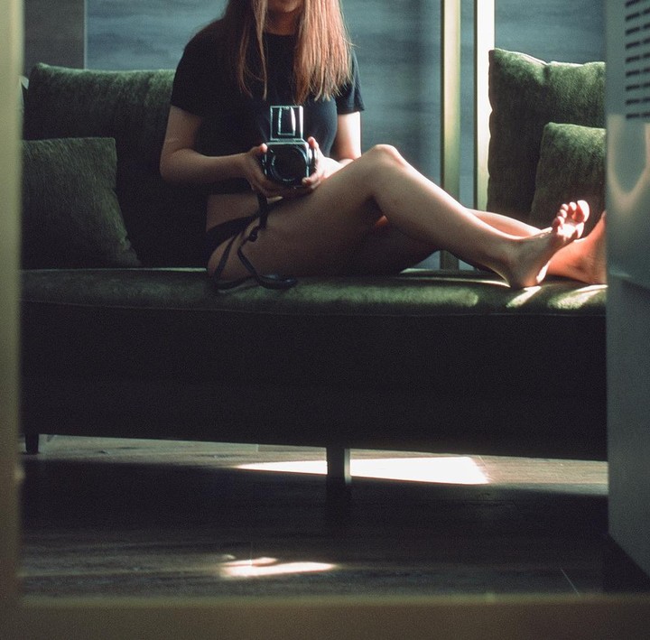 A woman sits on a green velvet couch, holding a camera, with sunlight illuminating her legs and feet.