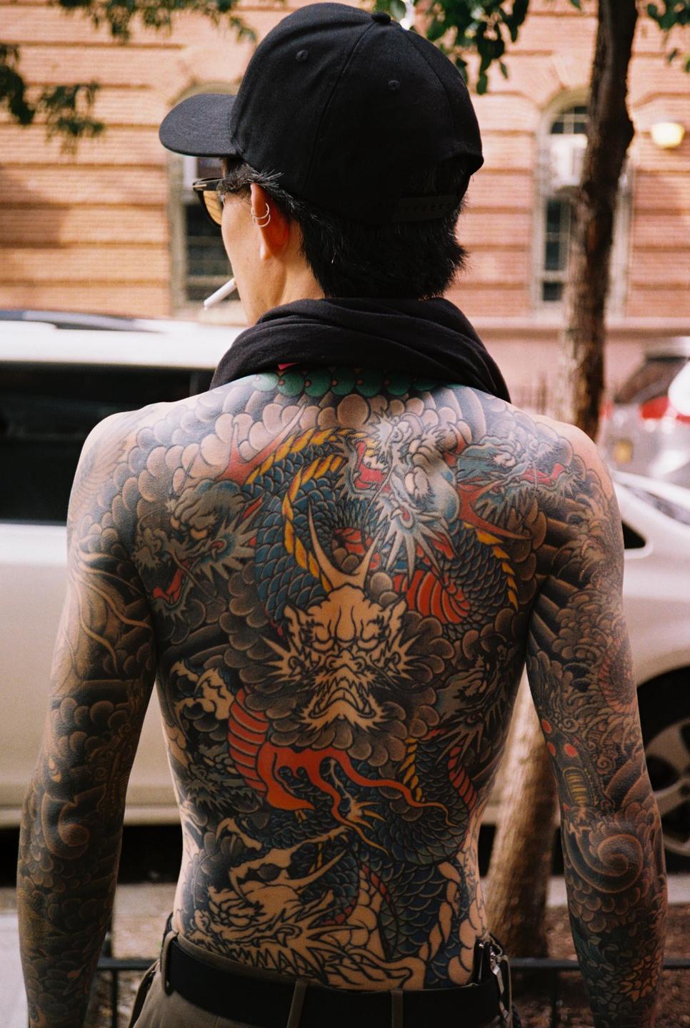 A man with a full back tattoo of dragons and clouds is seen from behind, wearing a black cap and smoking a cigarette.