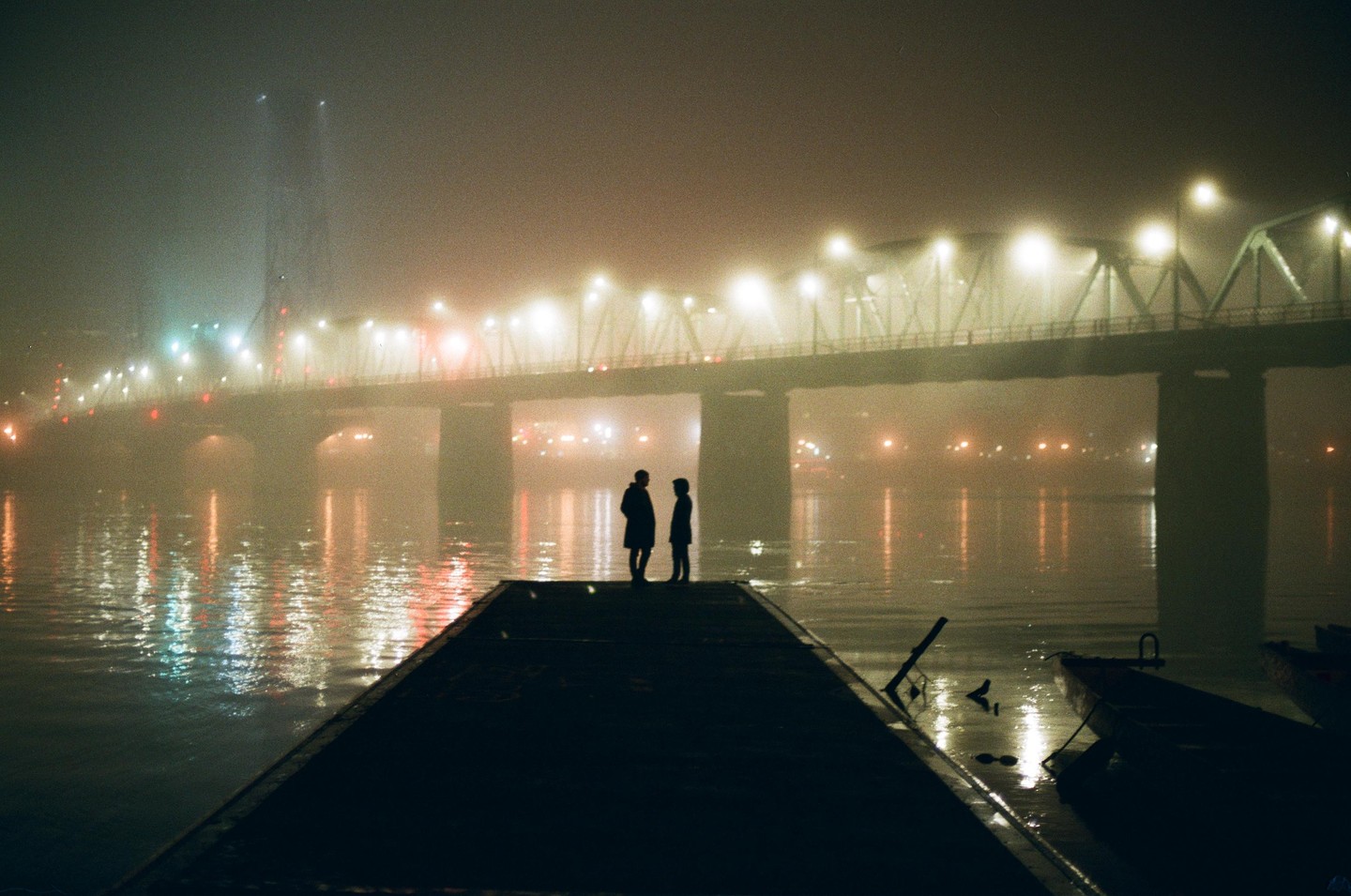 Two people stand on a dock at night with a bridge and its reflection in the water behind them.