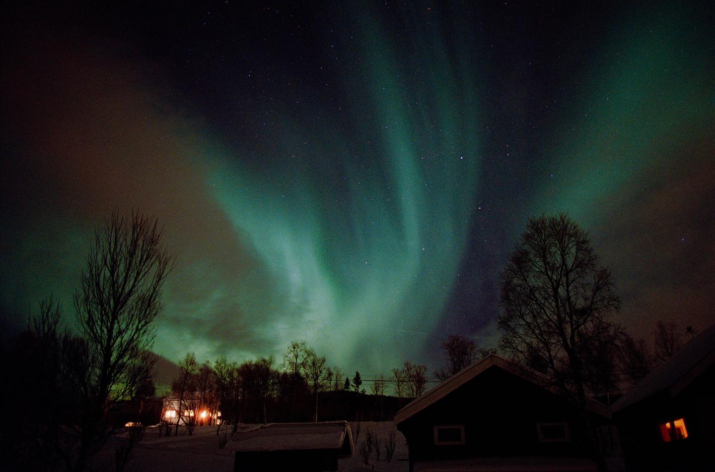 The aurora borealis illuminates the night sky above snow-covered houses and trees.