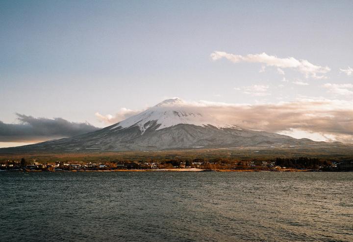Mount Fuji rises above a town and a body of water under a cloudy sky.