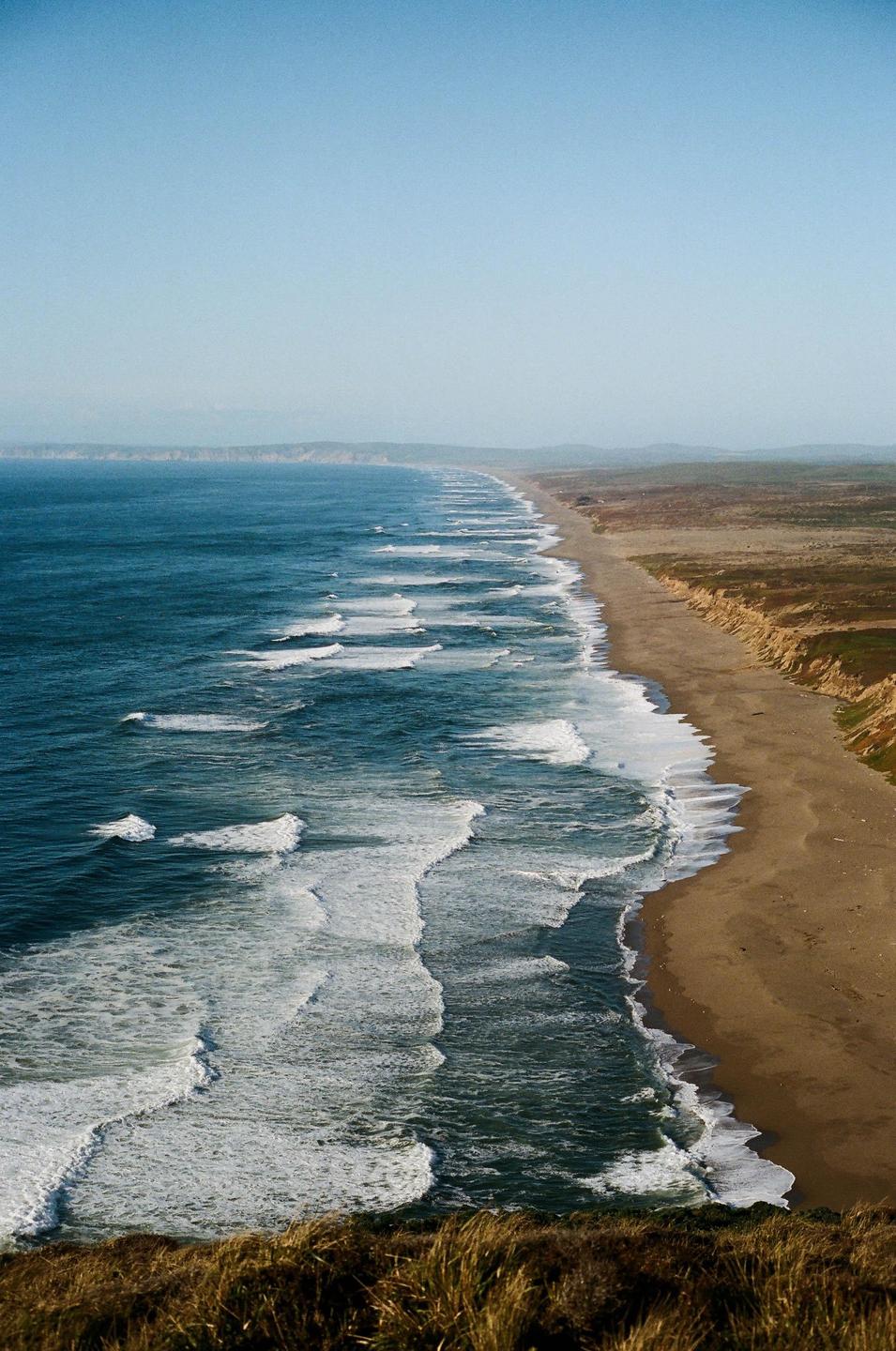 Waves crash on a long sandy beach next to a cliff under a clear sky.