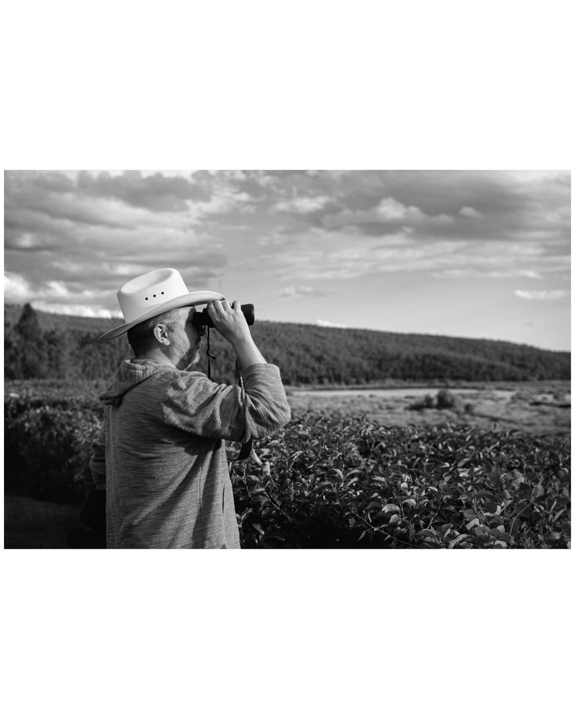 A man wearing a cowboy hat looks through binoculars at a distant landscape.