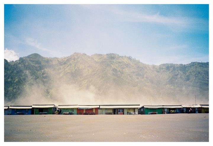 A row of small, colorful shacks sits on a sandy plain with a large, hazy mountain in the background.