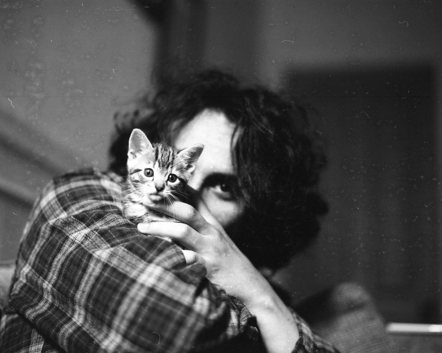 A person with dark hair and a plaid shirt holds a small kitten.