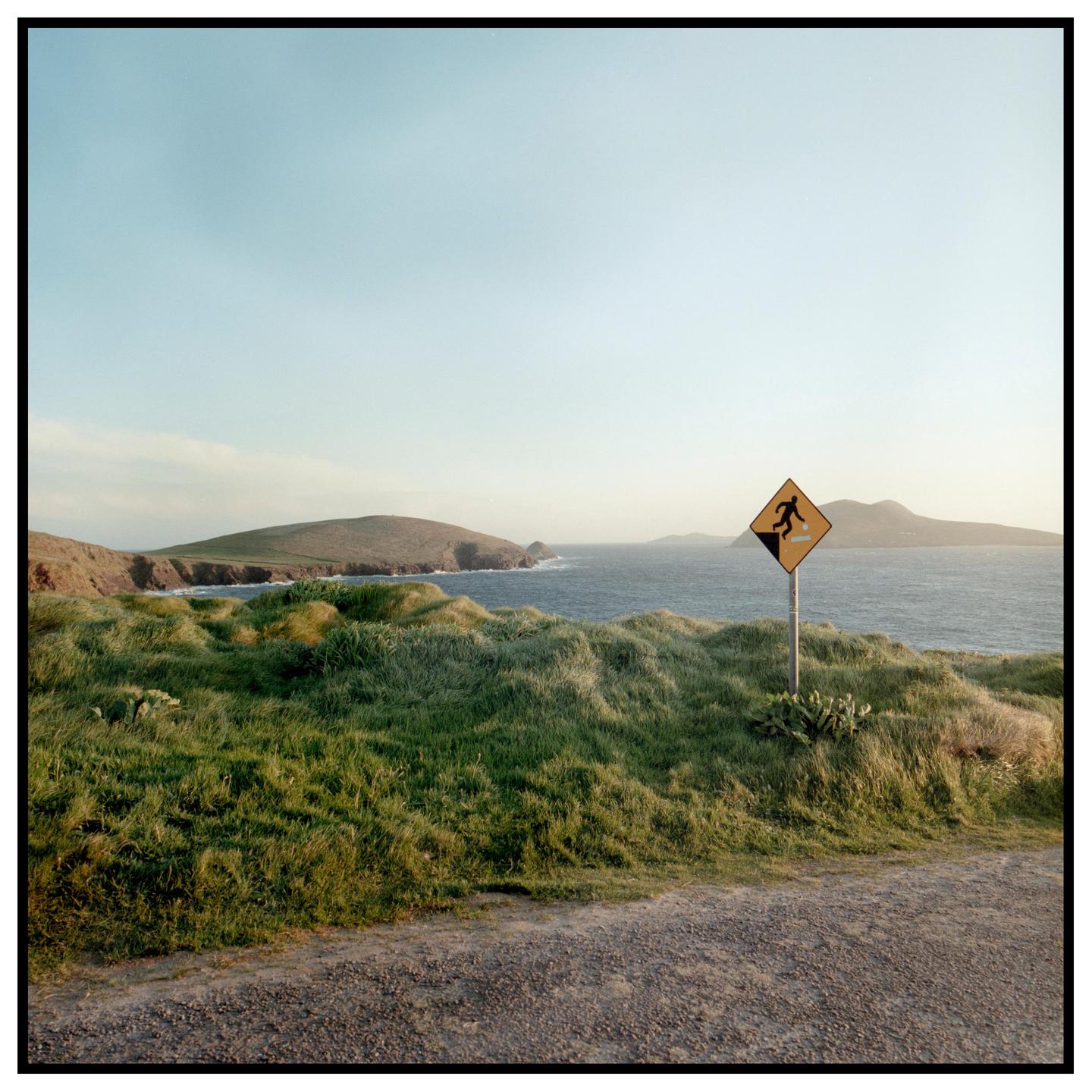 A warning sign for falling rocks stands on a grassy hill overlooking the ocean and distant landmasses.