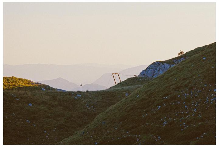 A mountain landscape shows a valley with a small structure and a distant mountain range under a clear sky.
