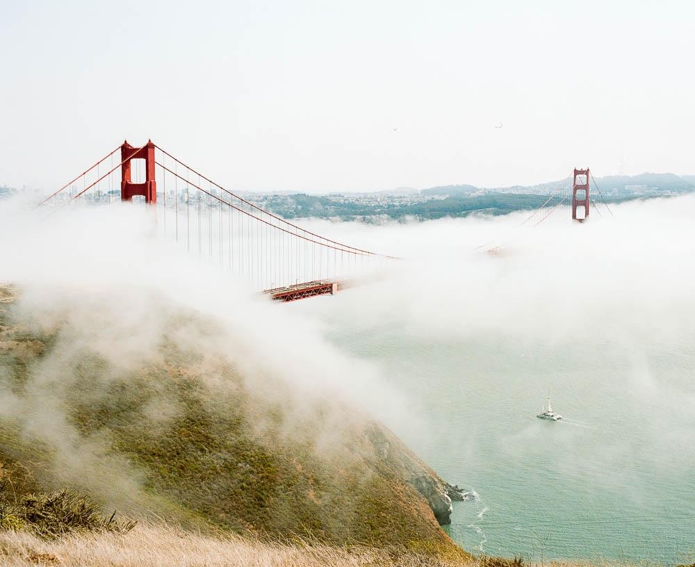 The Golden Gate Bridge is partially obscured by fog, with a sailboat visible on the water below and a grassy hillside in the foreground.