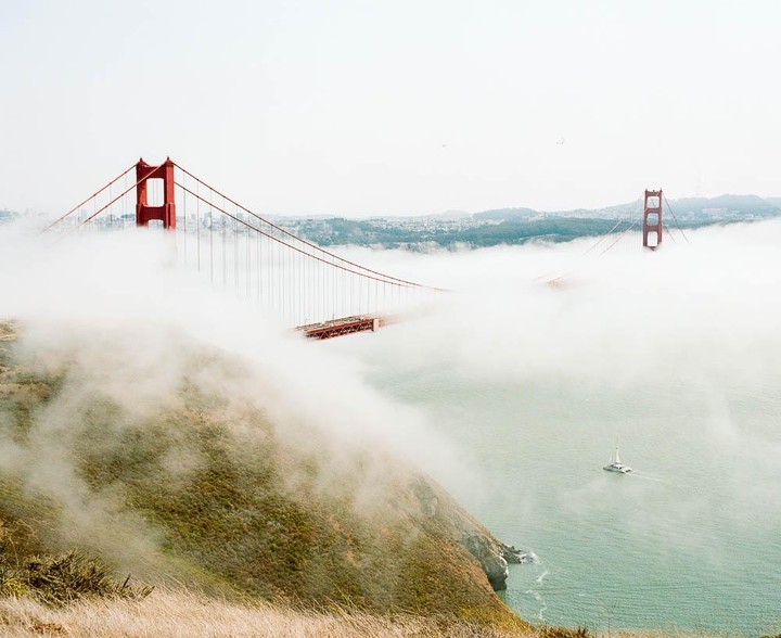 The Golden Gate Bridge is partially obscured by fog, with a sailboat visible on the water below and a grassy hillside in the foreground.