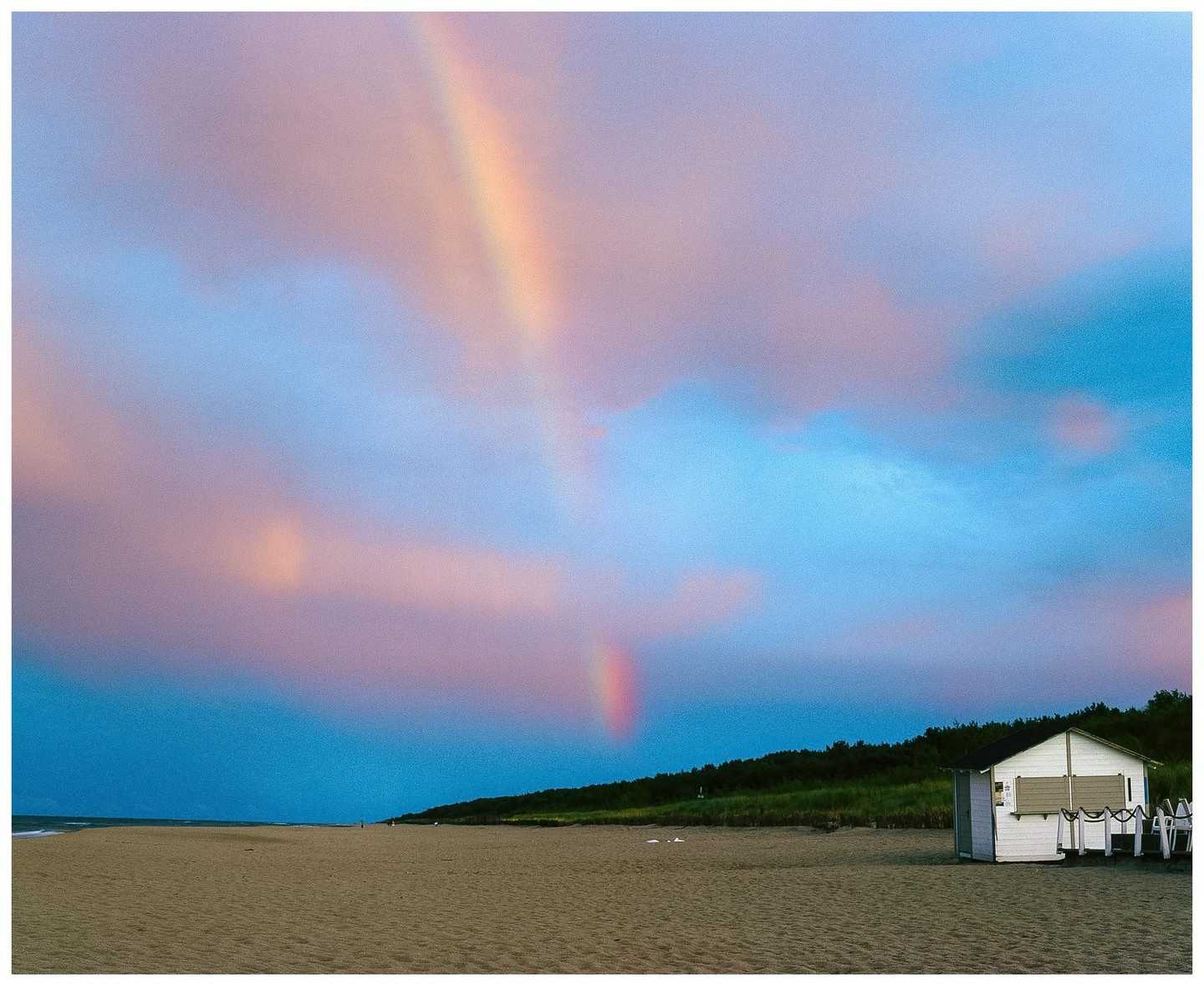 A rainbow is visible over a sandy beach with a small white building and a line of trees under a colorful sky.