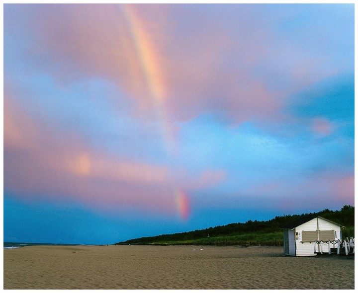 A rainbow is visible over a sandy beach with a small white building and a line of trees under a colorful sky.