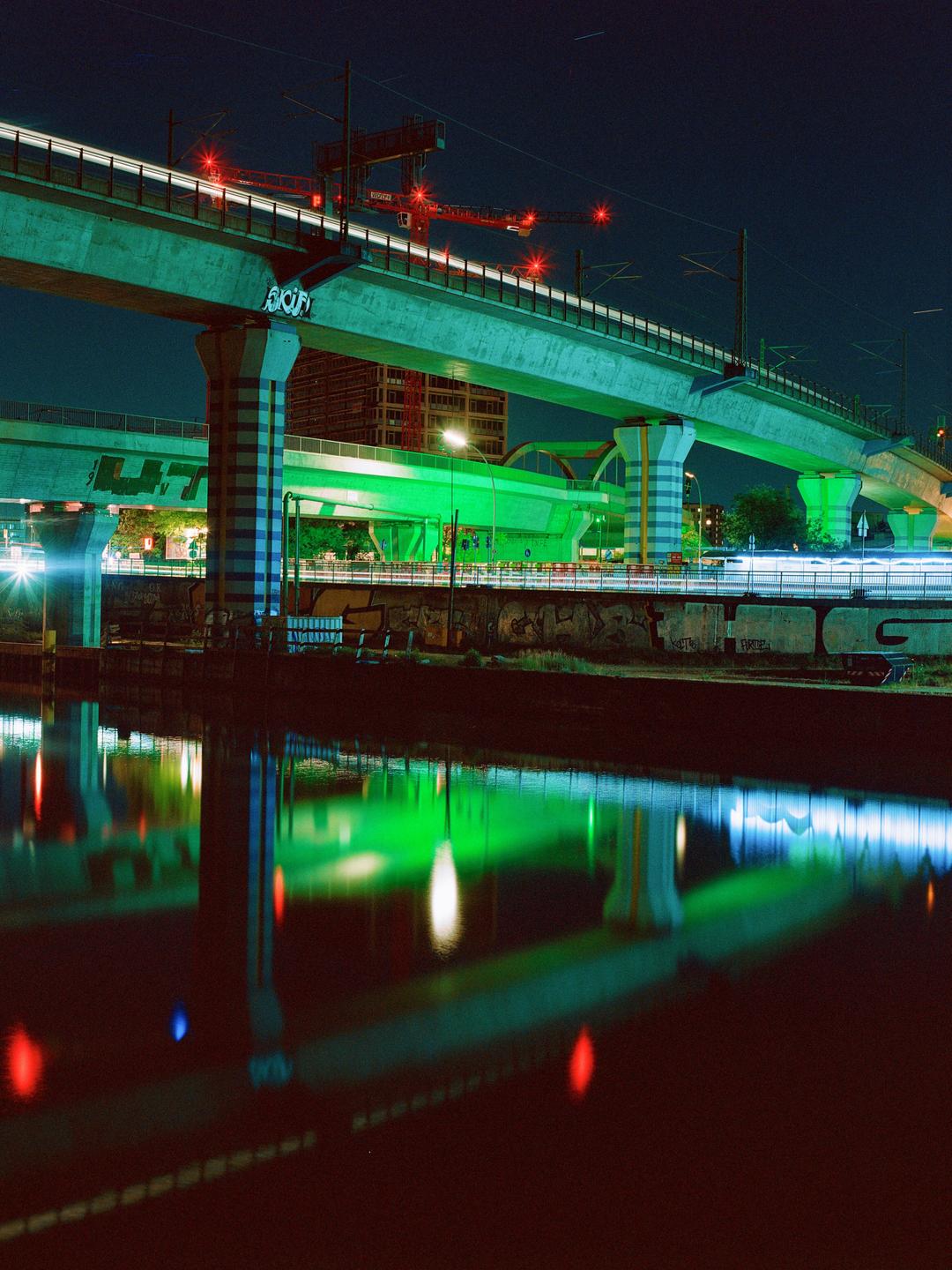 Light trails from vehicles on a bridge are reflected in the water below at night.