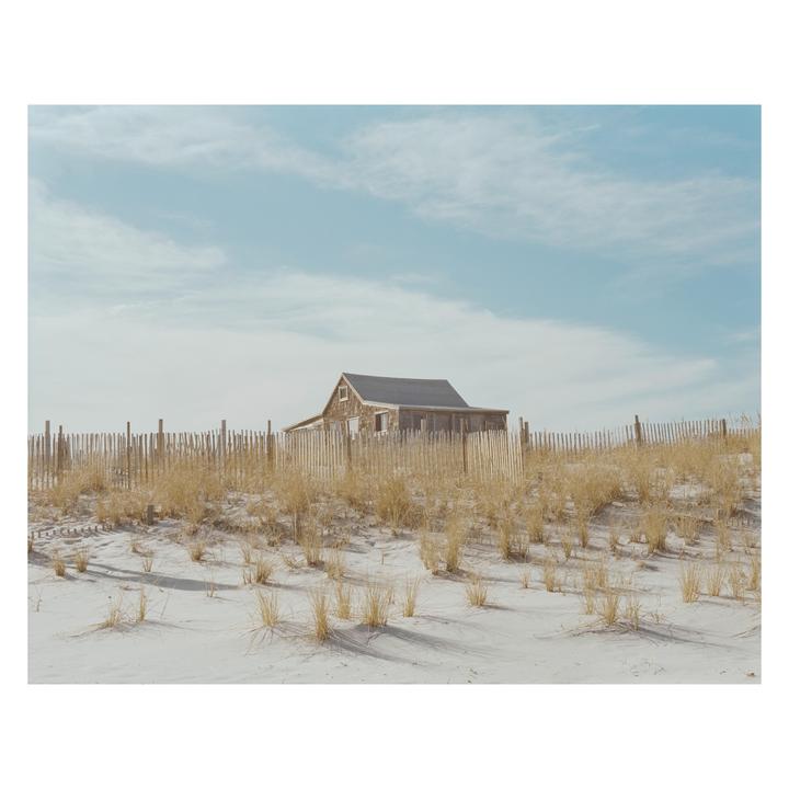 A weathered wooden shack stands behind a picket fence and dry grass on a sandy beach under a blue sky.