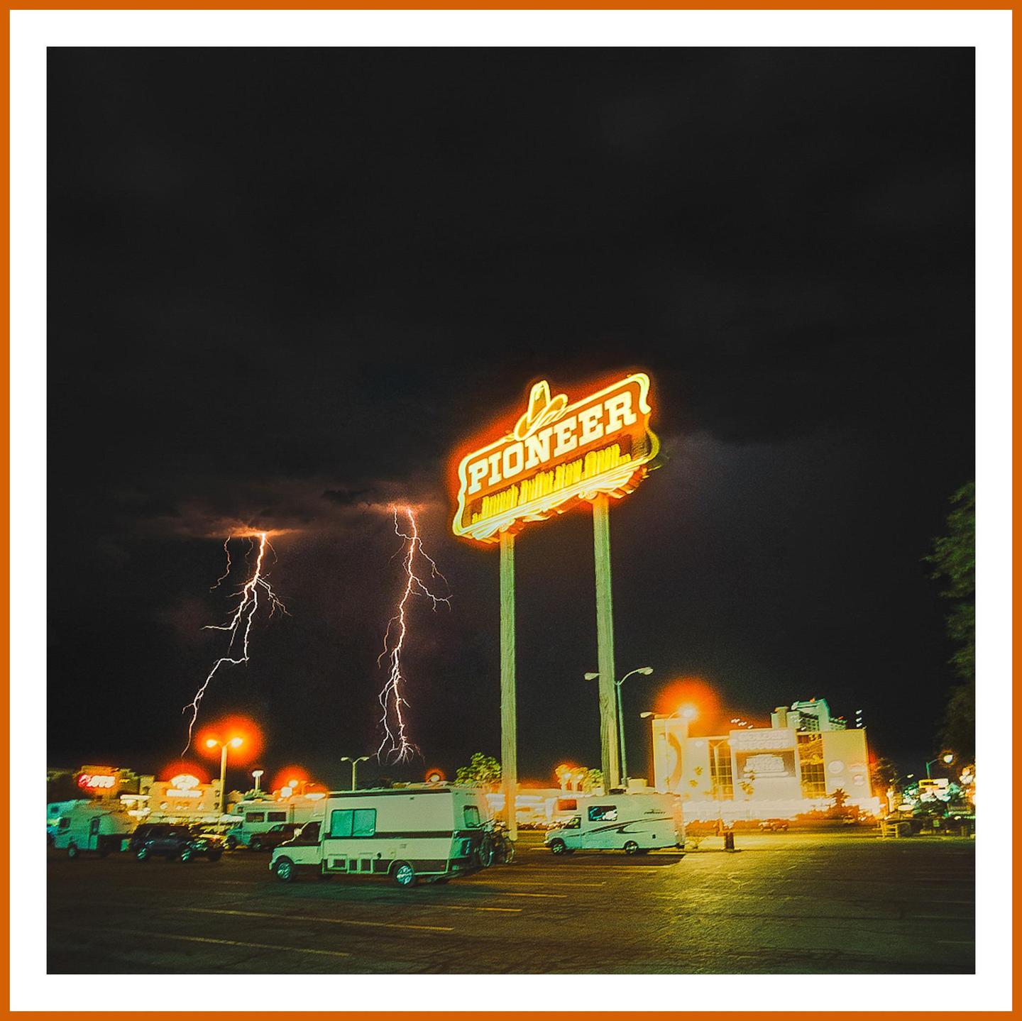 Lightning strikes behind a neon "Pioneer" sign in a parking lot at night.