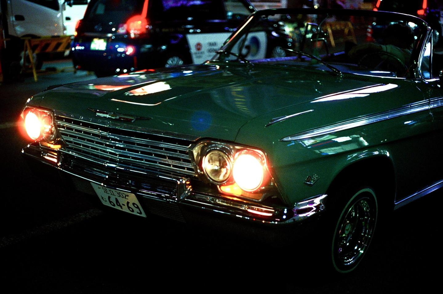 A green vintage car with its headlights on is parked on a street at night.