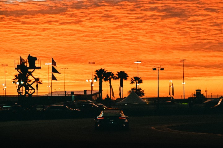 A car is visible on a track with palm trees and stadium lights silhouetted against an orange sunrise.