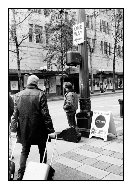 A man with a suitcase walks past a street musician and a "one way" sign on a city street.