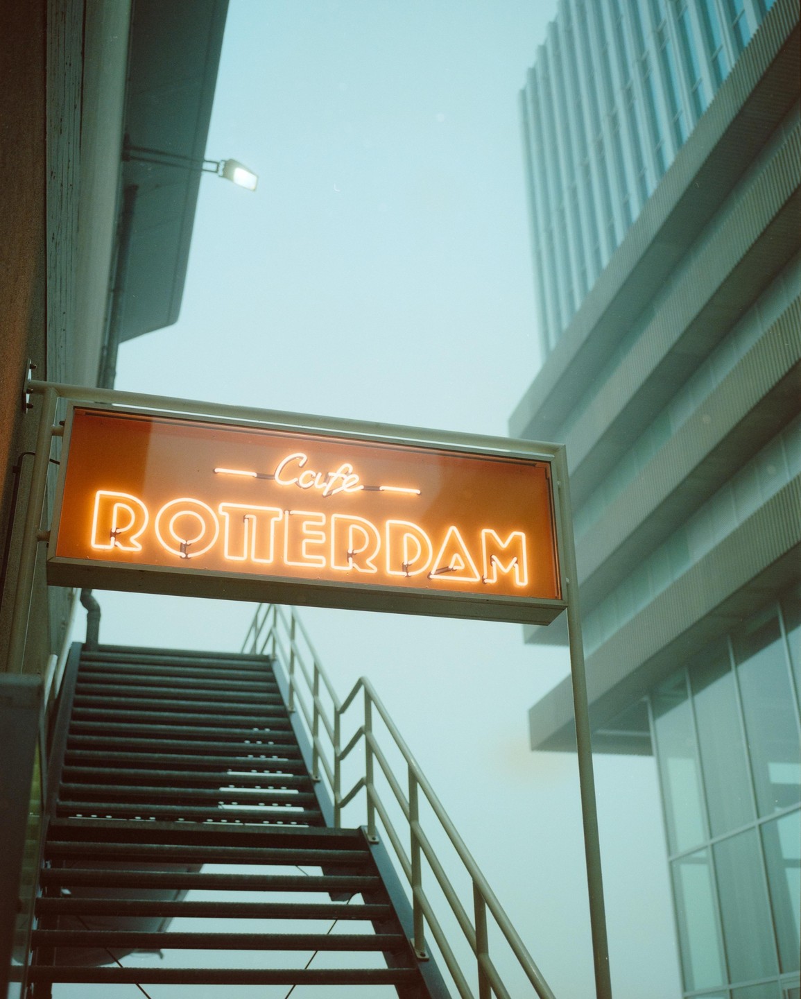 An orange neon sign for "Cafe Rotterdam" hangs above a metal staircase next to a modern building.