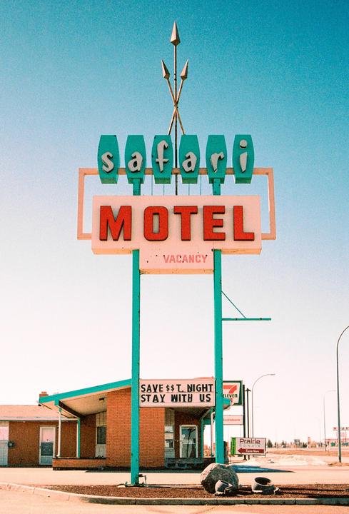 A large motel sign with the words "Safari Motel" and "Vacancy" stands in front of a brick building with a teal roof.