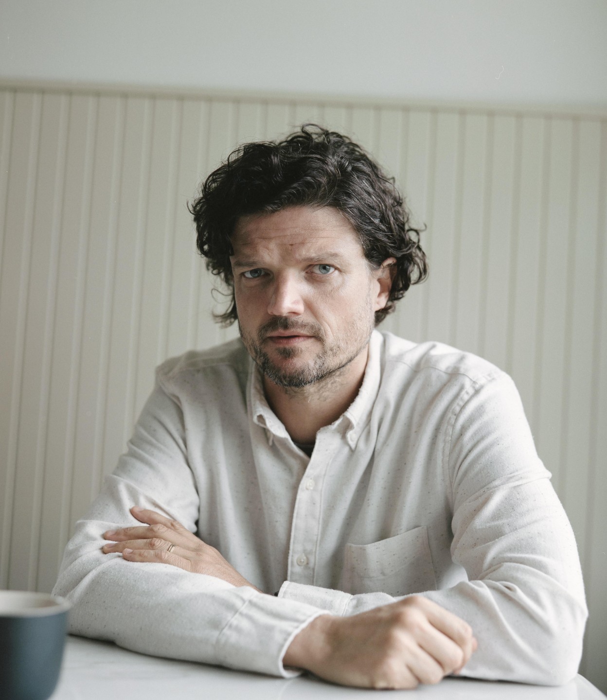 A man with curly hair and a beard sits at a table with his arms crossed.
