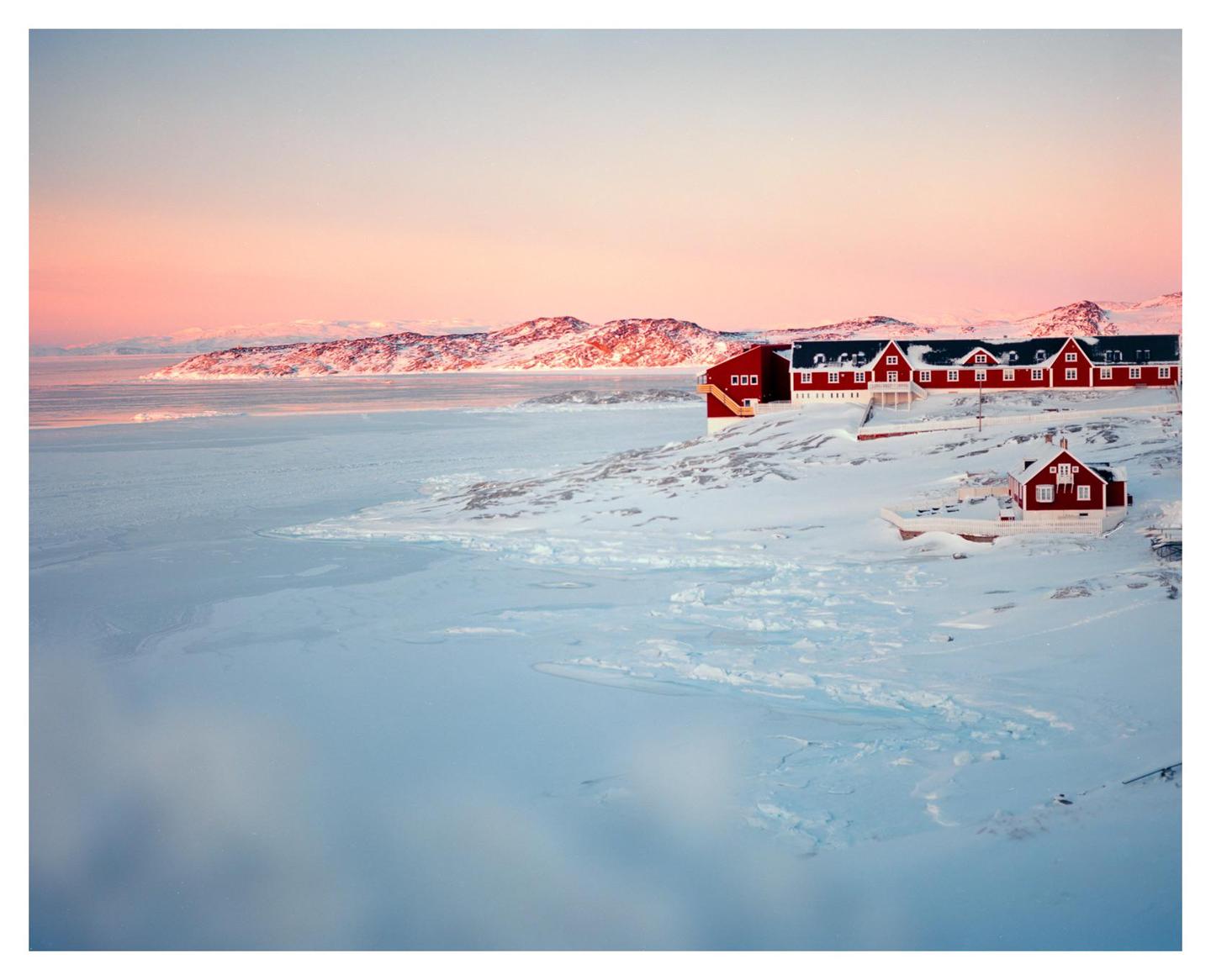 Red houses sit on a snow-covered landscape next to a frozen body of water with mountains in the distance under a pink sky.
