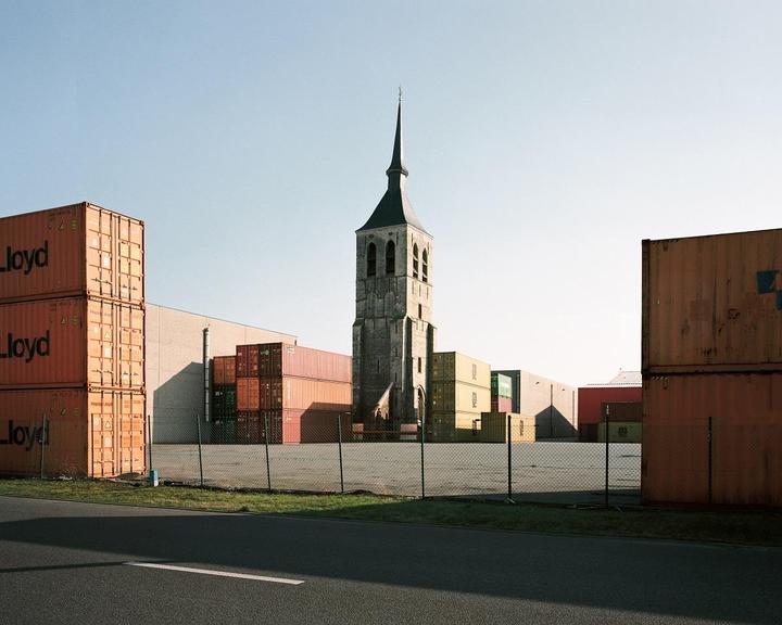 A tall church steeple rises above stacks of shipping containers in an industrial area.