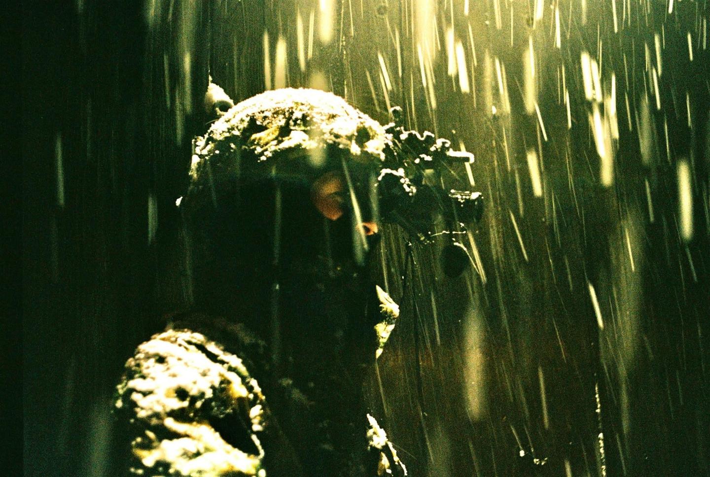 A soldier wearing a helmet and night vision goggles is seen from the side in heavy rain.