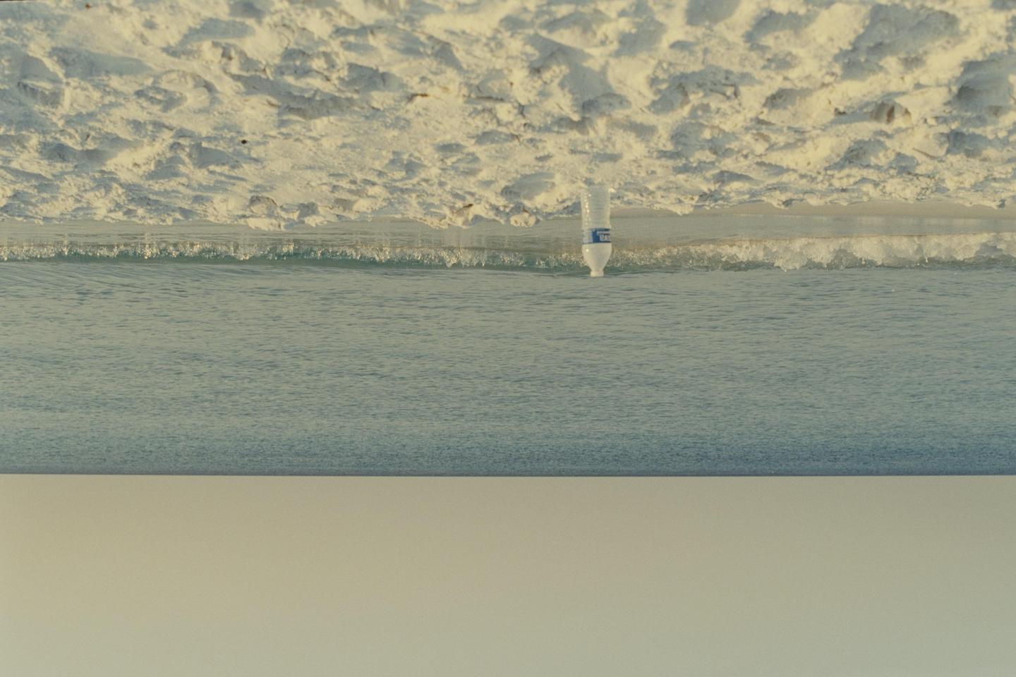 A plastic bottle floats upside down in the ocean near a sandy beach.