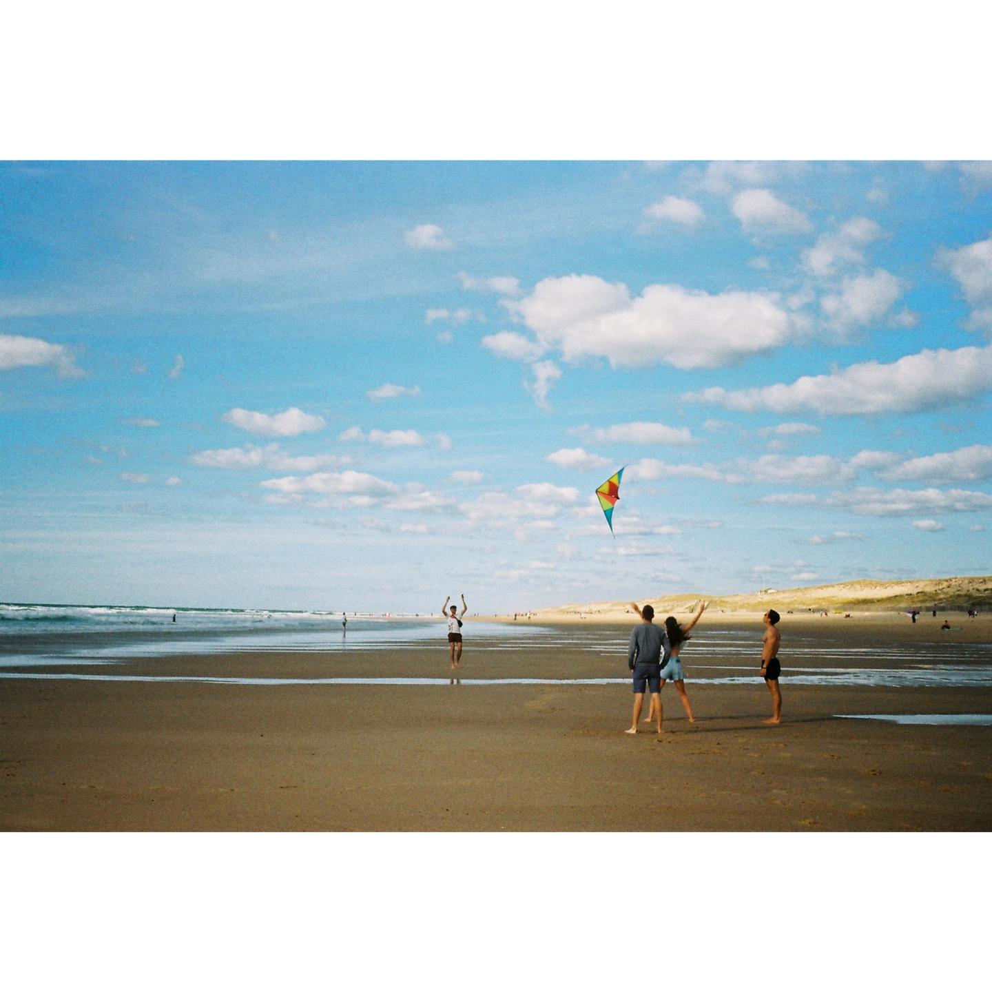 Three people stand on a beach looking at a kite flying in the sky while another person stands in the distance.