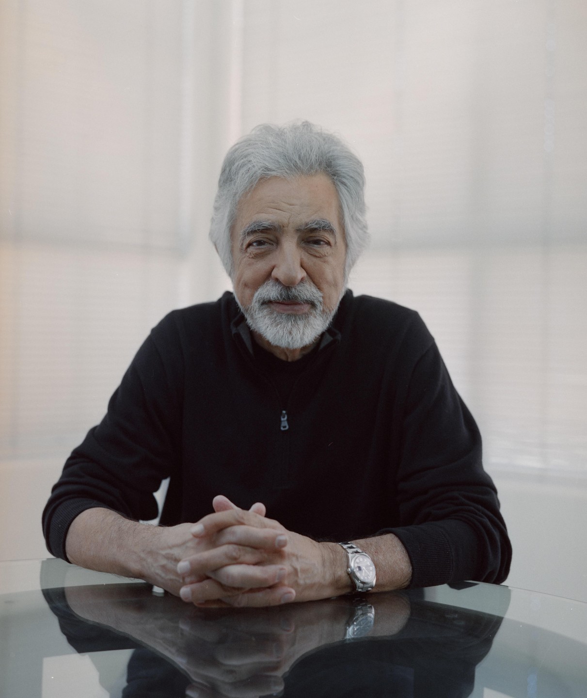 A man with gray hair and a beard sits at a table with his hands clasped.