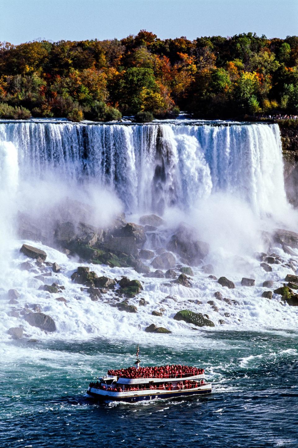 A boat full of people approaches a large waterfall with autumn trees on the cliff above.