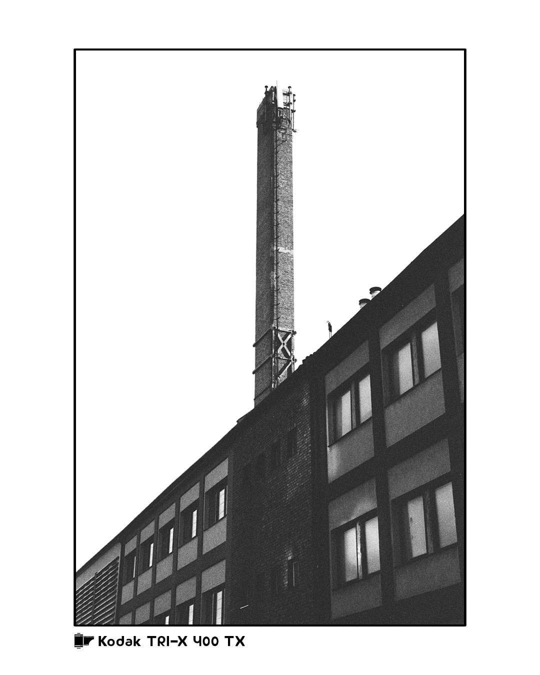 A tall, narrow chimney rises above a building with many windows.