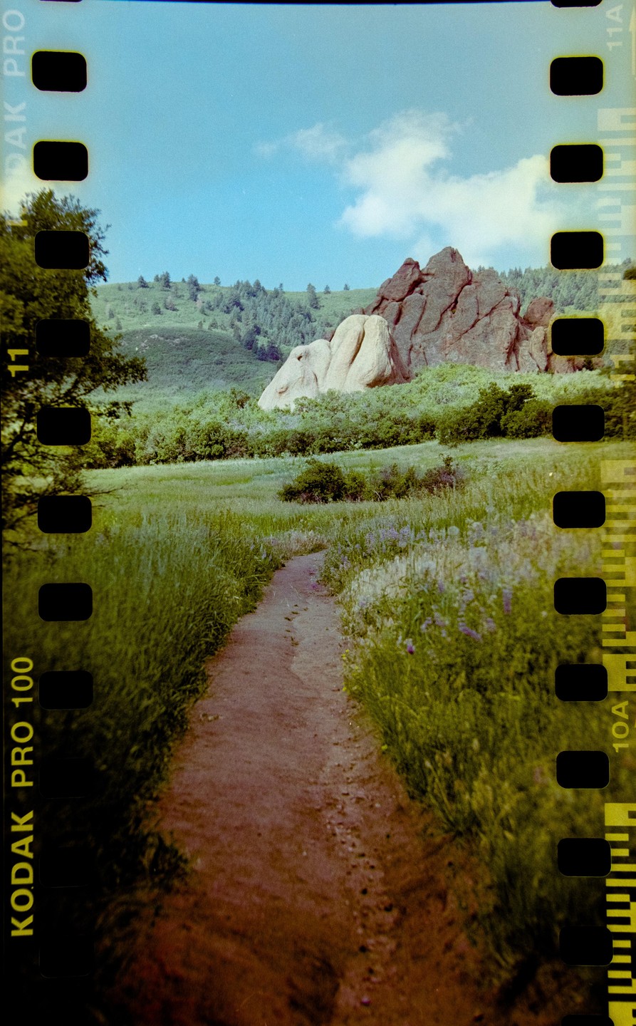 A dirt path leads through a grassy field towards large rock formations and a tree-covered hill under a blue sky.