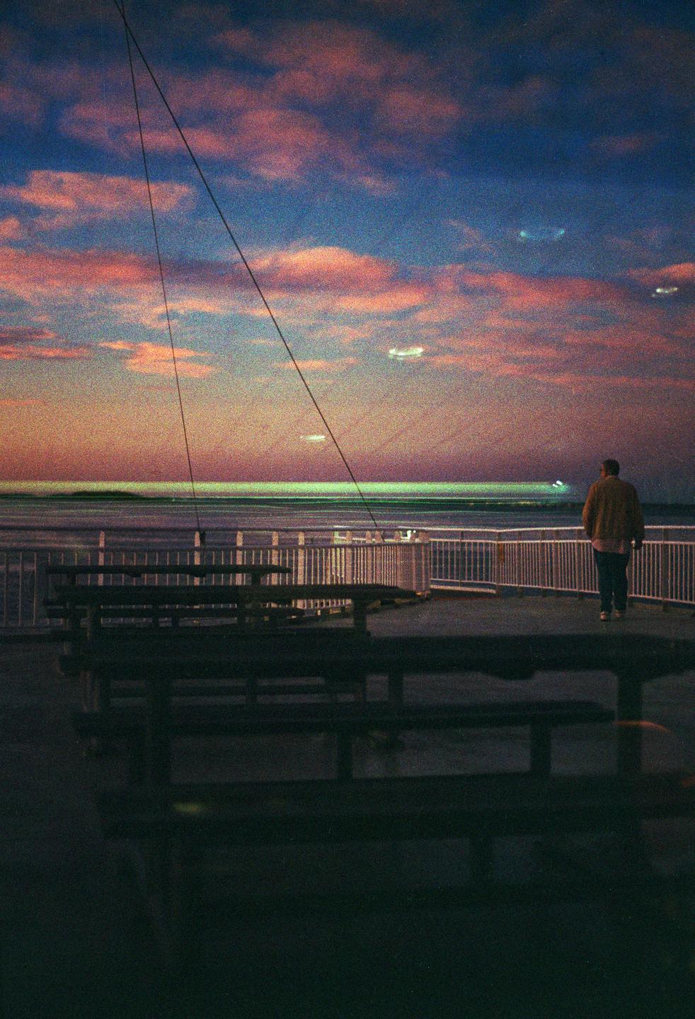A man stands on a ferry deck looking out at the ocean under a colorful sunrise sky.