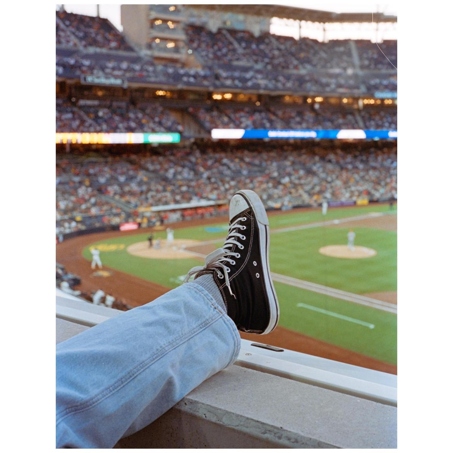 A person's leg with a black sneaker rests on a ledge overlooking a baseball stadium filled with spectators.