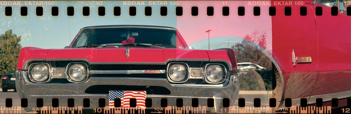 A red vintage car with a chrome bumper and an American flag license plate is parked outdoors.