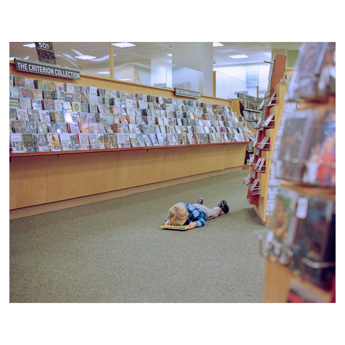 A child lies face down on a book on the carpeted floor of a bookstore.