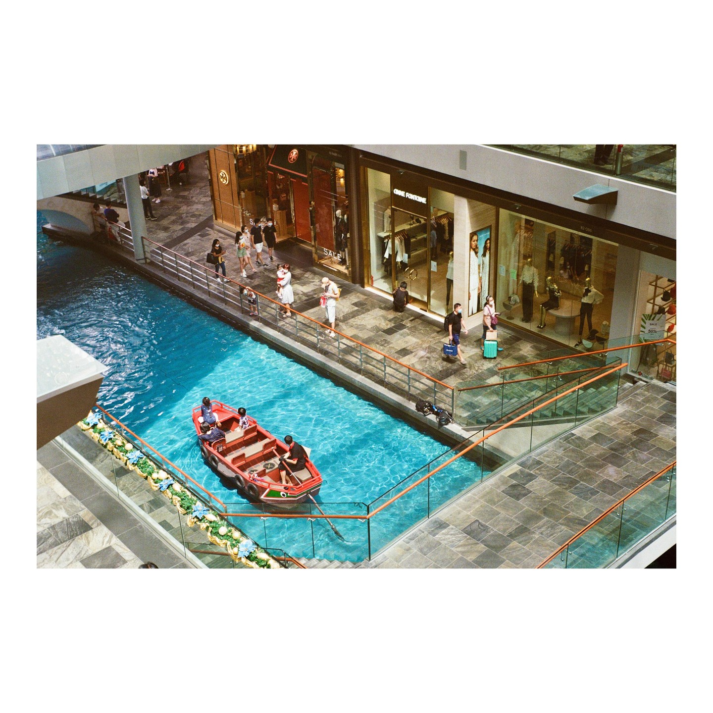 A red boat with people floats on a bright blue waterway inside a shopping mall with people walking along the banks.
