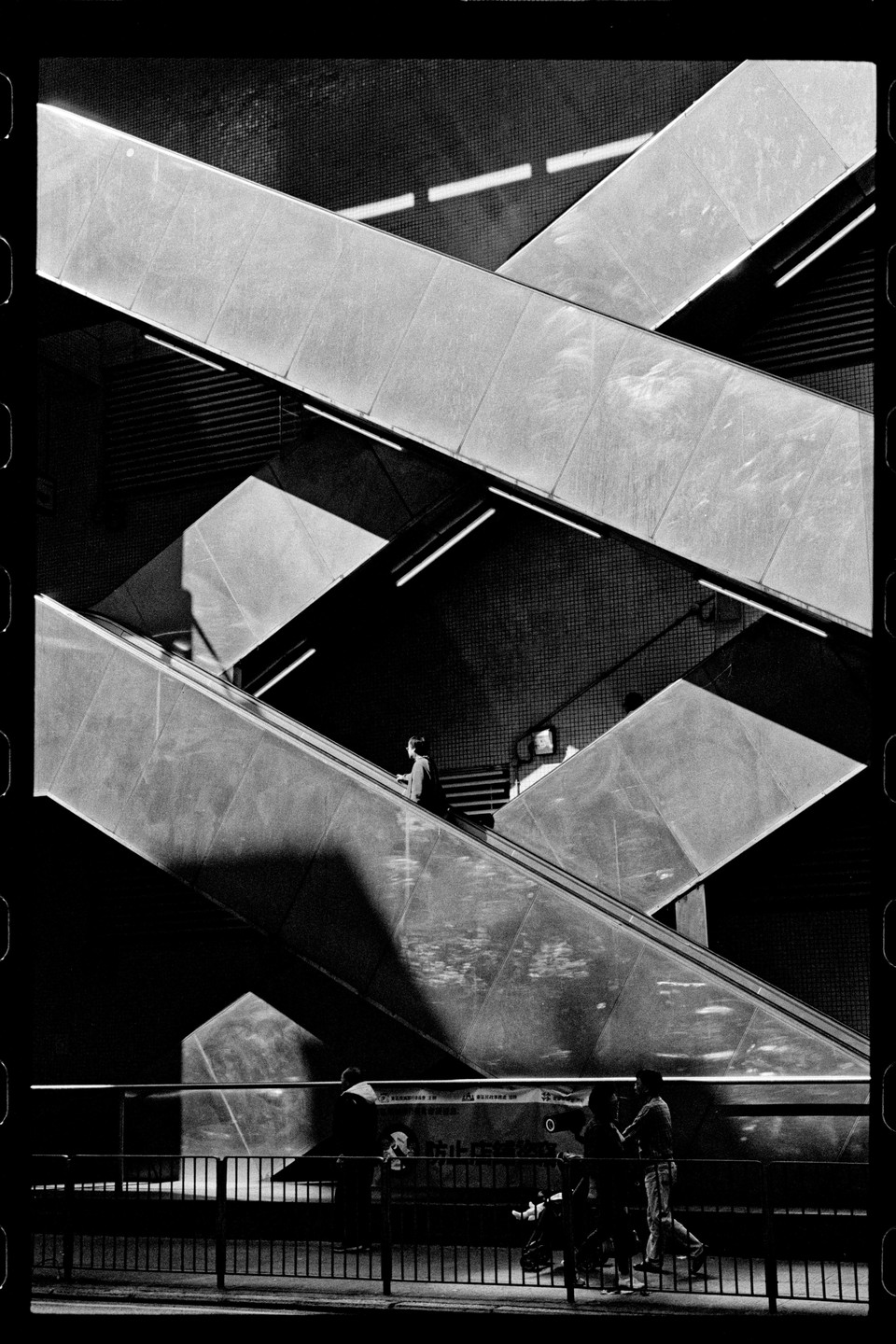 A person rides an escalator while other people stand on a street below a complex arrangement of escalators and stairs.