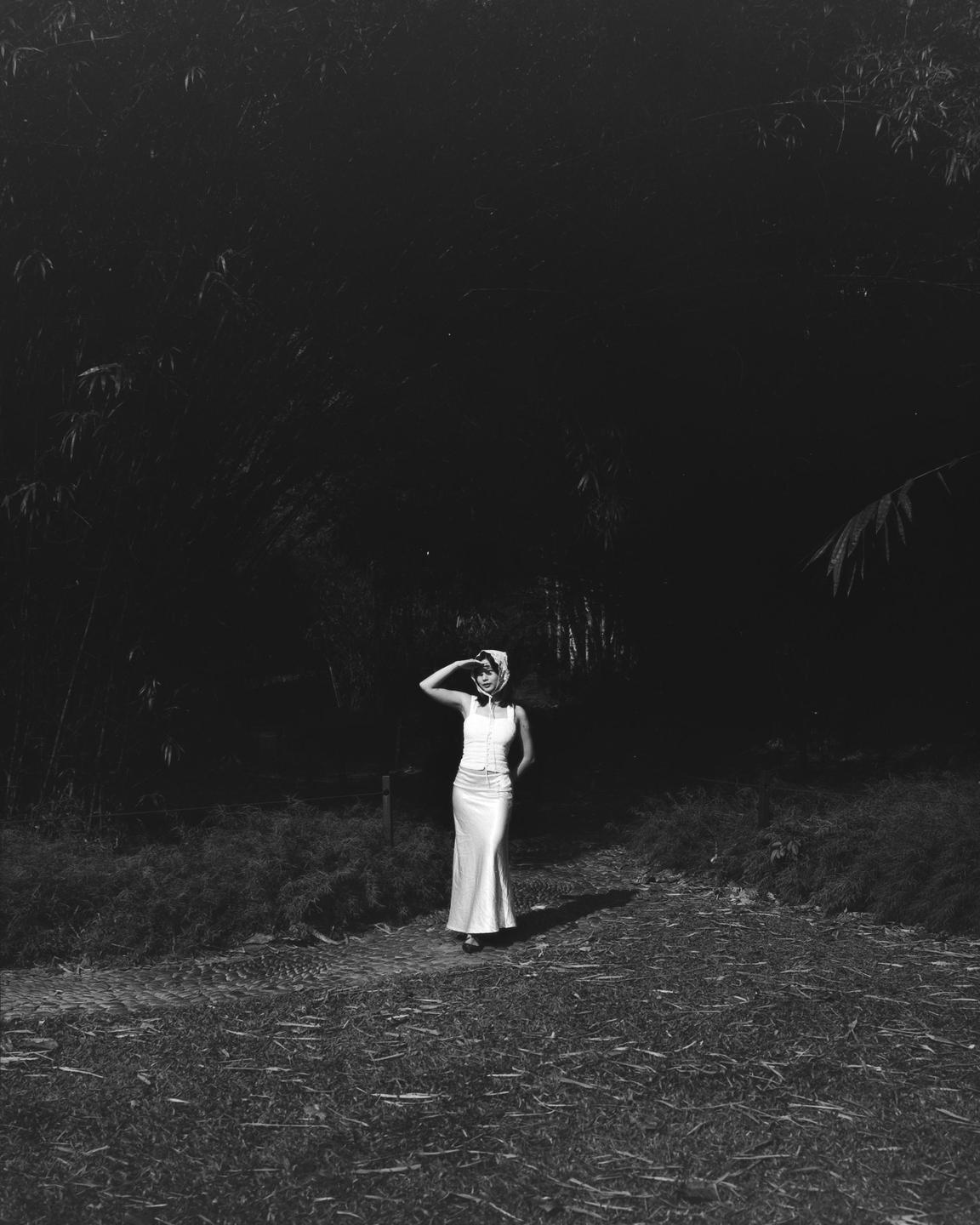 A woman in a white dress and headscarf stands on a path with dark foliage in the background.