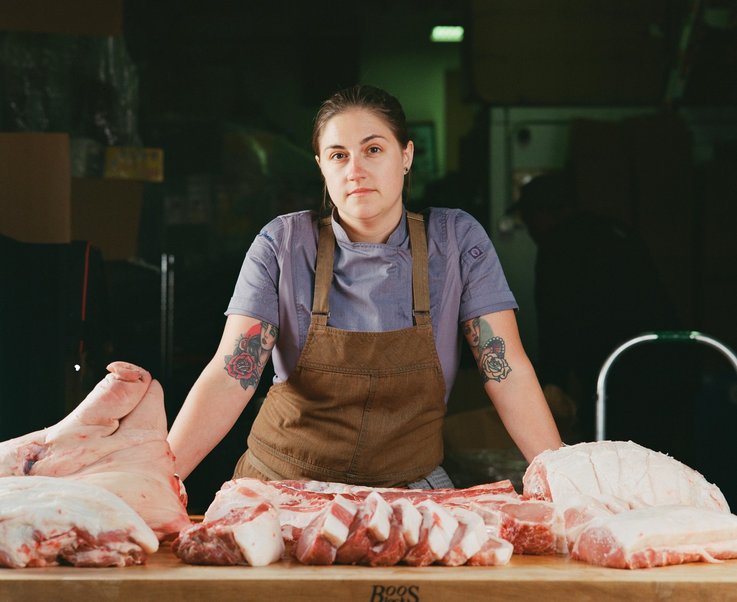 A woman in an apron stands behind a wooden counter with various cuts of pork.