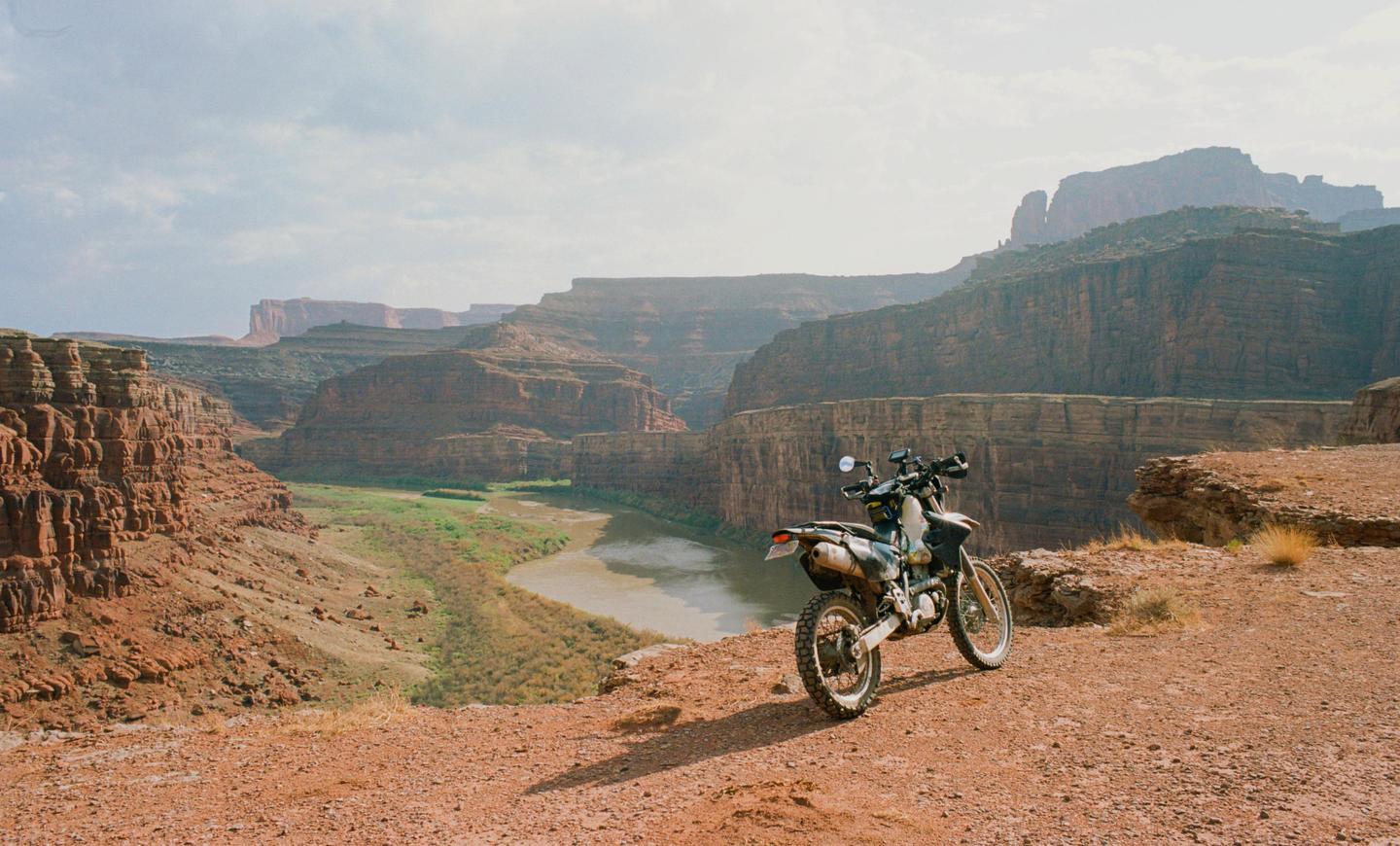 A motorcycle is parked on a dirt path overlooking a river winding through a canyon.