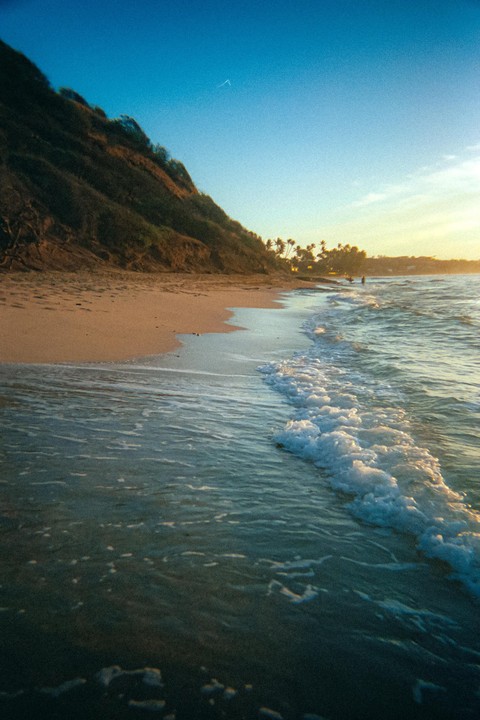 Waves crash on a sandy beach next to a hill covered in vegetation under a bright sky.