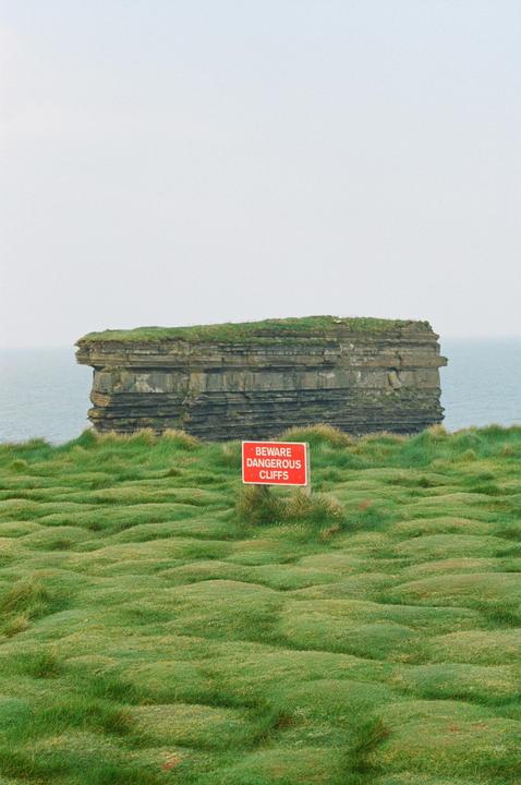 A red sign warning of dangerous cliffs stands in a field of green grass with a large rock formation in the ocean behind it.