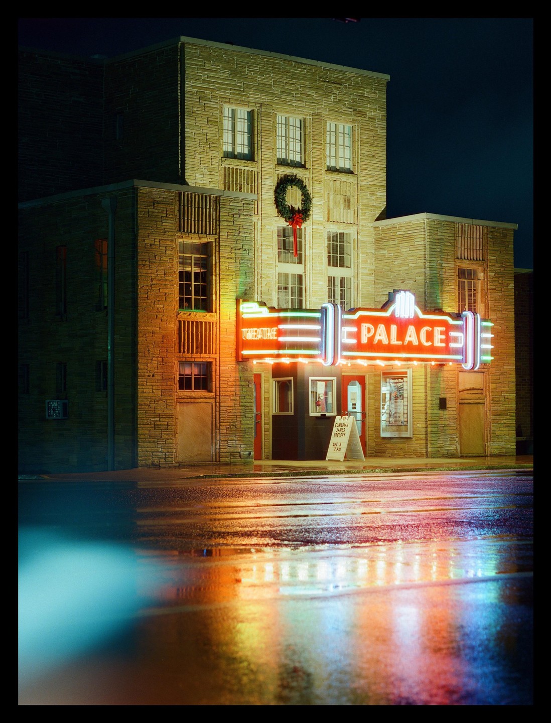 A building with a neon sign for "Palace" is reflected in a wet street at night.