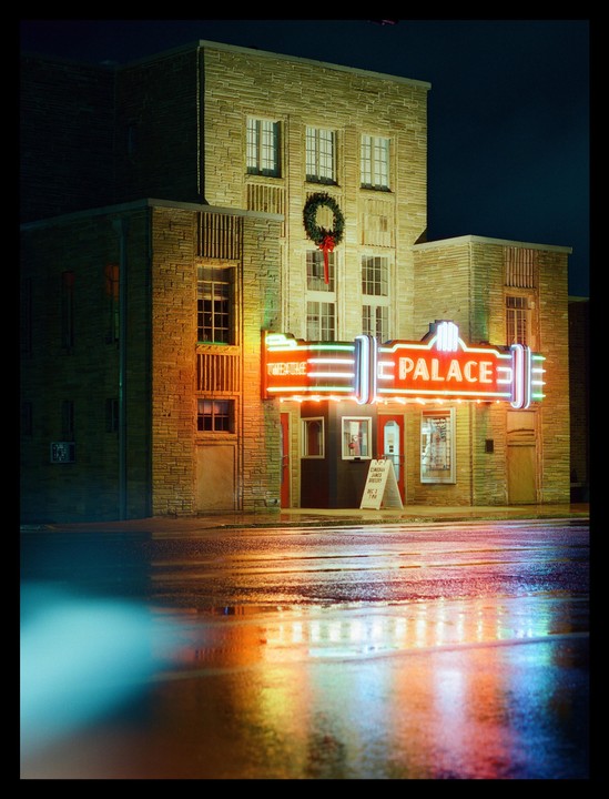 A building with a neon sign for "Palace" is reflected in a wet street at night.
